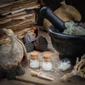 Bottles of homeopathic globules, mortar, dried moss, old books, dry roots, nuts and plants on table.