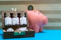 Bottles of homeopathic medicine in old wooden box and a coin on pink piggy bank with blue surface and wood background - saving Royalty Free Stock Photo