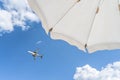 Bottom view on white beach or pool umbrella and blue sky with small cloudy and flying plane. Copy space. Holiday, vacation, travel