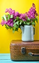 Bouquet of lilac in an old enameled teapot on vintage suitcase on yellow background. Retro style still life.