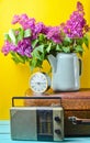 Bouquet of lilacs in enameled kettle on antique suitcase, vintage radio, alarm clock on yellow background. Retro style still life. Royalty Free Stock Photo