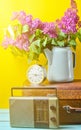 Bouquet of lilacs in enameled kettle on antique suitcase, vintage radio, alarm clock on yellow background. Retro style still life. Royalty Free Stock Photo