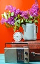 Bouquet of lilacs in enameled kettle on antique suitcase, vintage radio, alarm clock on yellow background. Retro style still life. Royalty Free Stock Photo