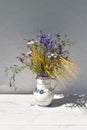 A bouquet of meadow wild flowers in a white jug on a wooden white table, in the background a white wall with a shadow Royalty Free Stock Photo