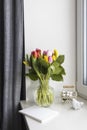 A bouquet of multi-colored tulips in a transparent vase on the windowsill on a holiday. White notebook, wicker metal basket with