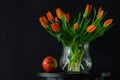 bouquet of orange tulips in a transparent round vase and an apple on a dark background Royalty Free Stock Photo