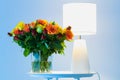 A bouquet of orange yellow and red roses flowers on a table next to a lamp with blue background close up view