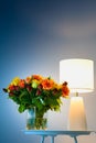 A bouquet of orange yellow and red roses flowers on a table next to a lamp with blue background close up view