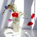 Bouquet of three red poppy flowers and different wildflowers in crystal vase with water on white table with contrast sun light and Royalty Free Stock Photo