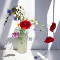 Bouquet of three red poppy flowers and different wildflowers in crystal vase with water on white table with contrast sun light and Royalty Free Stock Photo