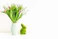 Bouquet of tulips in light green ceramic vase and Easter green grass bunny on white wall background.