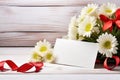 A bouquet of white daisies and a gift box on a table with red satin ribbons, a sheet of notes and red hearts on a Royalty Free Stock Photo