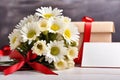 A bouquet of white daisies and a gift box on a table with red satin ribbons, a sheet of notes and red hearts on a Royalty Free Stock Photo