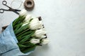 A bouquet of white tulips in blue wrapping paper in wooden box with scissors and twine on a white concrete background