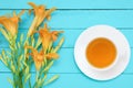 Bouquet of yellow flowers hemerocallis and white cup on saucer with green tea on a turquoise wooden background. View from abov Royalty Free Stock Photo