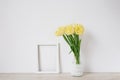 A bouquet of yellow spring tulips in a vase stands on the table, next to an empty white frame for a photo with copy space Royalty Free Stock Photo