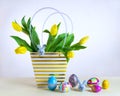 Bouquet of yellow tulips in paper striped bag. Group of Easter painted eggs and decorative funny bunnies on light yellow table