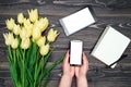 Bouquet of yellow tulips, tablet, open empty notebook and cellphone with white blank screen in woman hands on wooden background