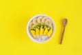 Bowl of oatmeal decorated with banana, kiwi, mango and coconut flakes and wooden spoon on yellow background.