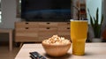 Bowl of popcorn, remote control and glass of beverage on wooden desk.