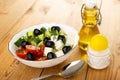 Bowl of salad with cucumbers and tomato, black olives, cheese, bottle of vegetable oil, salt, spoon on wooden table