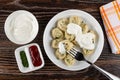 Bowl with sour cream, sauce boat with ketchup and parsley, fork in plate with dumplings, napkin on wooden table. Top view