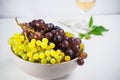 Bowl of various grapes: red, white and black berries on the white wooden table with glass of white wine and green leaf in the back