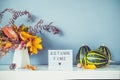 Box with text AUTUMN TIME, decorative striped pumpkins, vase with bouquet of falling leaves and fern on gray blue wall background