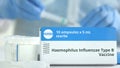 Box with haemophilus influenzae type B vaccine on the table against blurred lab assistant. Fictional phaceutical logo