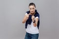 Boxing. Adult woman in casual clothes with blue jeans and sweater on her shoulders, smart watch on arm, ready for fight on grey b