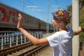 A boy in a beautiful colored hat with a smile waves to a train passing by on the railway, seeing off or meeting passengers