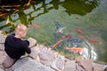 Boy in black clothes is on the shore of the rocks and feeds the koi carp in the pond Royalty Free Stock Photo