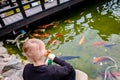 Boy in black clothes is on the shore of the rocks and feeds the koi carp in the pond Royalty Free Stock Photo