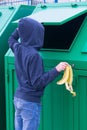 A boy in a blue hooded jumper throws a banana skin into a green dumpster