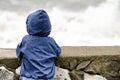 Boy in blue jacket with hood stands with his back against the pier against the background of sea waves Royalty Free Stock Photo