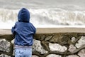 Boy in blue jacket with hood stands with his back against the pier against the background of sea waves. Royalty Free Stock Photo