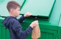 A boy in a blue jumper, throws a bottle in a paper bag into a green dumpster