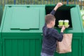 A boy in a blue jumper throws empty disposable glasses and a paper bag into a green dumpster Royalty Free Stock Photo