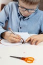 Boy in a blue shirt and glasses sits at a desk and concentrates on doing plastic work with colored paper. The boy is painting with