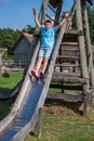 A boy in a blue t-shirt and denim shorts slides down a wooden playground slide, arms raised with exhilaration. Perfect