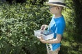 Boy in a blue T-shirt and a straw hat on his head stands near the bush of Kamchatka berries. He holds in his hands a container w