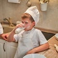 A boy in chef clothes drinks milk from a glass while cooking an apple pie in the kitchen Royalty Free Stock Photo