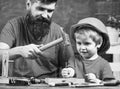 Boy, child busy in protective helmet learning to use hammer with dad. Father with beard teaching little son to use tools