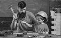 Boy, child busy in protective helmet learning to use hammer with dad. Father with beard teaching little son to use tools