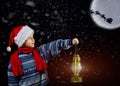 Boy in Christmas hat with lantern in hand, points the way Santa Claus flying on his sleigh with the moon Royalty Free Stock Photo