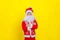 A boy in a .d santaclaus suit and an artificial beard stands on a yellow background and crosses his arms on his chest