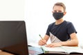 A boy in a disposable mask at home in front of a computer does his homework. Distance learning in quarantine. Textbooks are on the Royalty Free Stock Photo