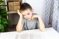 The boy doesn`t want to read the book. A boy in a gray T-shirt sits at a white table at the open book and holds his hand behind