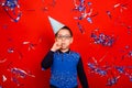 A boy in a festive cap blows into a festive pipe and stands in the rain of blue and silver confetti on a red background. Royalty Free Stock Photo