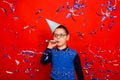 A boy in a festive cap blows into a festive pipe and stands in the rain of blue and silver confetti on a red background. Royalty Free Stock Photo